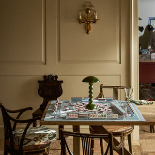 A mahjong game set arranged on a wooden dining table in a classic room with cream paneled walls, vintage wooden chairs, and a brass wall sconce.