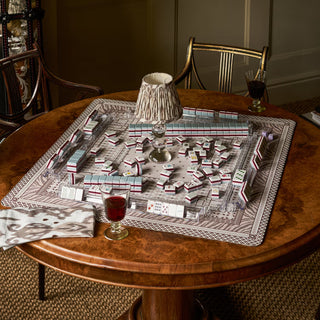 A mahjong game set arranged on a round wooden dining table in a real indoor room, with chairs, a table lamp, and two drinking glasses visible. The product appears to be the mahjong board and tiles, not the surrounding decor.