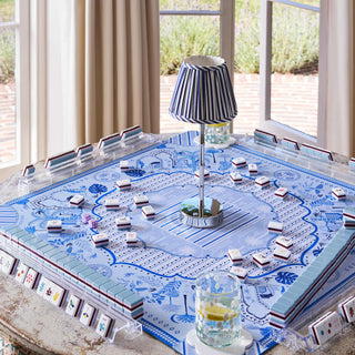A blue and white patterned board game set arranged on a table in a bright room with large windows, curtains, and an outdoor view. The set includes a rectangular game board with decorative blue illustrations, many small rectangular tiles, clear plastic storage bins, and a striped lamp in the center.