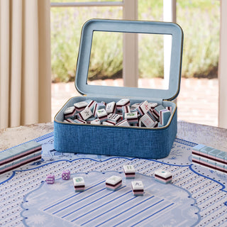 A blue fabric-covered mahjong set with an open hinged lid showing white tiles inside, placed on a patterned game mat on a table with a softly blurred indoor outdoor background.