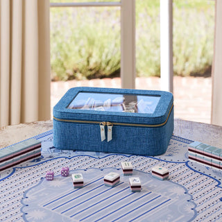 A blue fabric mahjong case with a clear top window sits on a patterned game mat on a table, with several tiles and dice arranged around it in a real indoor setting with a window and curtains in the background.