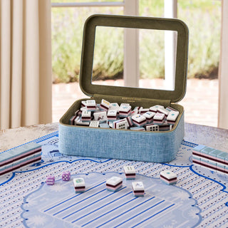 A blue fabric-covered mahjong set displayed open on a patterned game mat, with the lid propped up to reveal a clear lid insert and many white tiles with red and blue accents inside. A few tiles and dice are placed on the mat in front, and a real indoor setting with curtains and a window is softly visible in the background.
