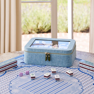A blue fabric-covered mahjong game set sits on a table in a bright room, with several tiles placed in front and a matching blue game mat on the surface. No person or animal is present, but the real indoor environment and window view make this Lifestyle rather than Product/Packaging.