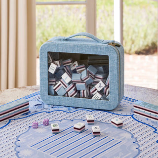A light blue fabric-covered mahjong set with a zippered carrying case, shown on a tabletop with tiles and dice in front. The background is a softly blurred indoor setting with curtains and a window.