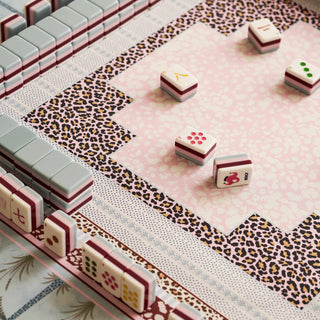 Mahjong game set with white tiles featuring red borders and various symbols, arranged on a pink patterned mat with leopard print borders.