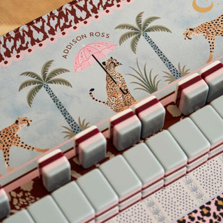 Close-up of a notebook or journal cover featuring a safari-themed illustration with cheetahs, palm trees, and a pink umbrella, overlaid with a keyboard-style row of pale gray and pink keys. The text 'ADDISON ROSS' is visible on the cover.