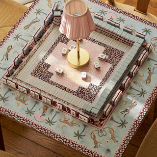 A square decorative mahjong-style game board displayed on a table in a real indoor setting, with a pink pleated table lamp placed in the center. The board has a pale blue border with animal and palm motifs, gray-green tiles along the edges, and a central play area with patterned tiles and small white dice.