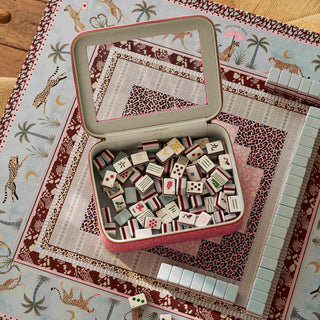 A pink metal tin of Mahjong tiles shown open on a decorative safari-print game mat, with several tiles scattered around it.
