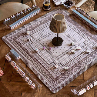 A square decorative table mat with an intricate geometric pattern in beige, brown, and gray, styled on a wooden table with a small table lamp, playing cards, and dice.