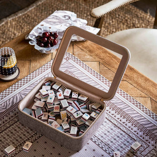 A square fabric storage box with an open hinged lid, filled with small rectangular game pieces, placed on a patterned runner over a wooden table. A glass candle holder, cherries, and part of a chair are visible in the styled setting.