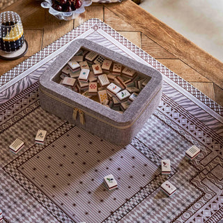A square gray fabric-covered mahjong storage box with a clear top, filled with wooden mahjong tiles, placed on a patterned rug over a wooden table.