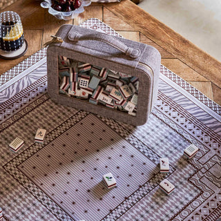A gray fabric game box filled with mahjong tiles sits on a patterned rug on a wooden table, with a few tiles placed outside the box.
