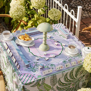 A styled outdoor dining table covered with a decorative tablecloth featuring a pastel game-board print, white teacups, plates, and a pale green table lamp, with flowers and a white fence in the background.