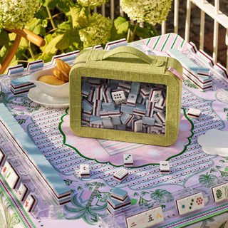 A light olive green fabric lunch box with a clear front window filled with miniature mahjong tiles, styled on a decorated board game table outdoors with greenery in the background.