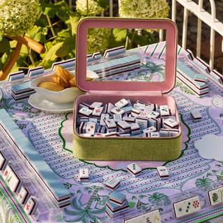 A pink and green Mahjong set displayed outdoors on a patterned table mat with a bowl of snacks nearby. The open rectangular tin contains the game tiles, with additional tiles arranged around it on a decorative surface.