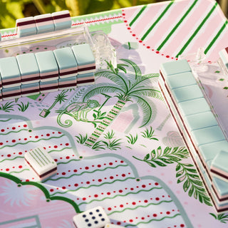 A close-up of a tropical-themed mahjong game set on a white table. The set includes mint green, white, and dark red tiles arranged on a decorative board printed with palm trees, green leaves, and red dotted patterns.
