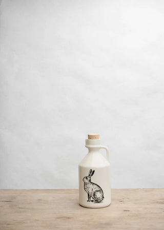 An off-white ceramic jug with a cork stopper and a black line-art illustration of a rabbit on the front, sitting on a light wood surface against a textured white wall.