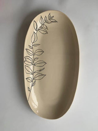 An oval ceramic serving platter in a warm cream-beige color, decorated with a delicate black line-art botanical leaf pattern running down the left side, set against a neutral gray background.