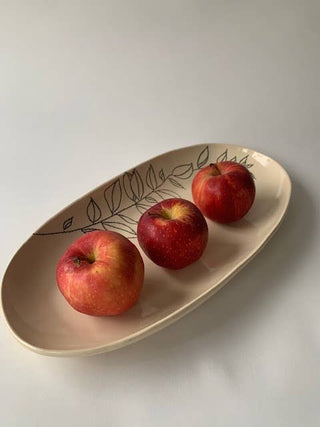 An oval beige ceramic serving platter with a black leaf pattern, holding three red apples on a light gray background.