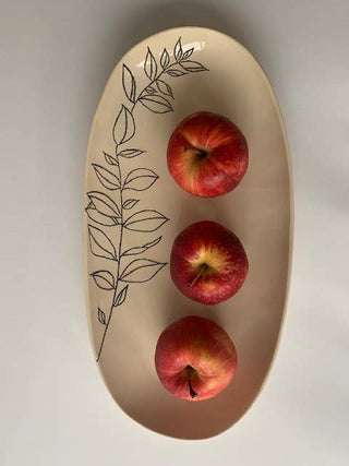An oval beige ceramic serving platter with a black line-art leaf design on one side, holding three red apples on a light gray background.