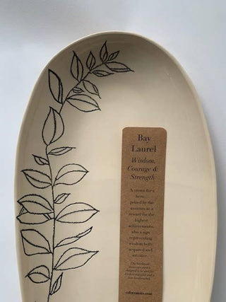An oval ceramic bowl in a warm cream color, decorated with a black line-art botanical leaf pattern on the left side and a brown paper label in the center.
