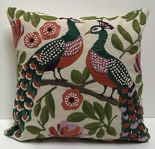 A square decorative throw pillow featuring an embroidered peacock design on an off-white background. The peacocks have dark green heads and bodies with rust-orange underbellies, pink patterned wings, and long green tail feathers.