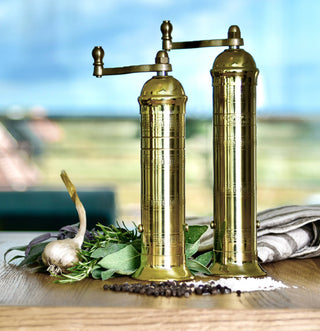 Solid brass pepper grinder with a textured finish, featuring a crank on top, set against a kitchen backdrop with herbs and spices.