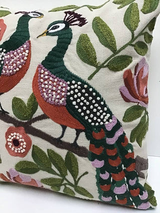 A square decorative throw pillow made of 100% cotton featuring an embroidered peacock and floral pattern on a cream fabric background.