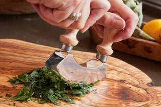 Person using a manual herb chopper on fresh greens on a wooden cutting board.