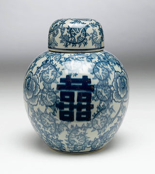 A photograph of a round ginger jar with a double happiness symbol and blue floral motifs on an off-white porcelain surface.
