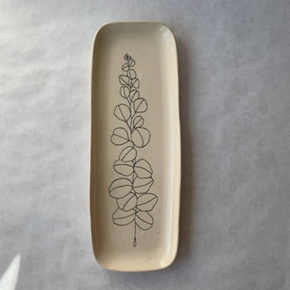 A rectangular ceramic dish with rounded corners, featuring a minimalist black line drawing of a leafy plant stem printed on a warm cream-colored glaze, set against a textured gray background.