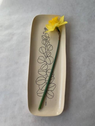 A rectangular beige ceramic plate with rounded corners, featuring a minimalist black line drawing of a leafy vine. A single yellow daffodil flower with a green stem is placed diagonally across the plate, which is set against a light gray textured background.