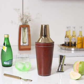 Brown leather cocktail shaker with gold accents on a kitchen counter with bottles and a glass.