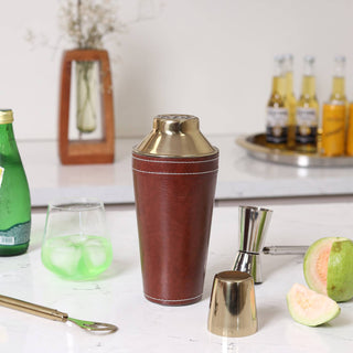 Brown leather cocktail shaker with gold accents on a kitchen counter with bottles and fruit.