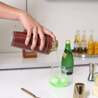 Person pouring a drink from a brown leather shaker into a glass with ice on a kitchen counter.