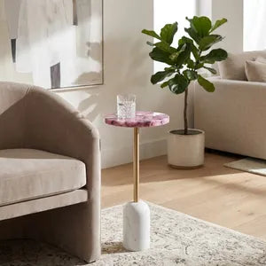 A modern accent side table in a living room setting, featuring a pink and white marble top, a gold metal stem, and a white cylindrical base.