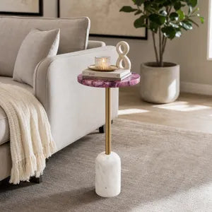 A modern side table with a purple and pink marbled top, a gold metal stem, and a white marble base, styled in a living room next to a sofa.