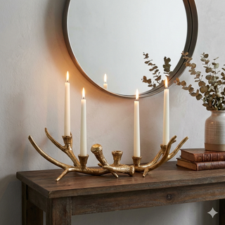 A gold-toned antler-style candle holder with four white taper candles, displayed on a rustic wooden console table in a styled interior setting with a round mirror, books, and a vase.