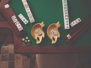 A top-down view of a mahjong game setup on a green felt table, featuring two gold fish-shaped dish holders containing dice, several tiles laid out, and wooden racks holding more tiles.
