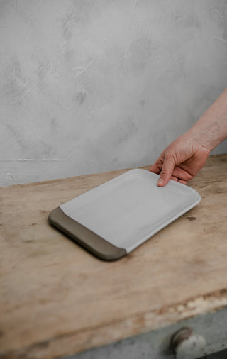A hand holding a rectangular white cutting board with dark brown silicone corners, resting on a rustic wooden table against a textured gray wall.