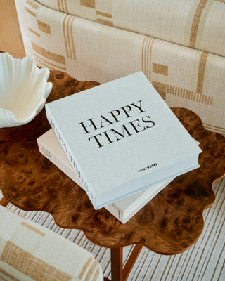 A light gray hardcover book titled 'HAPPY TIMES' with 'FASHIONWEAR' printed on the cover, resting on a scalloped wooden side table in a styled interior setting.