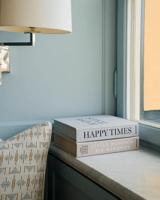 Two hardcover books, 'Happy Times' and 'Little Moments Big Memories', stacked on a light marble windowsill next to a patterned armchair and a wall-mounted lamp.