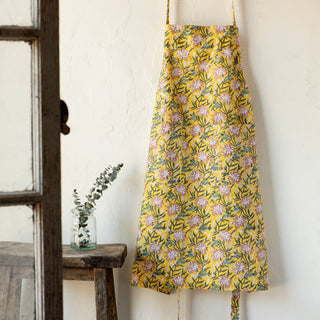 Yellow floral apron hanging on a white wall with a wooden stool and plant in the background.