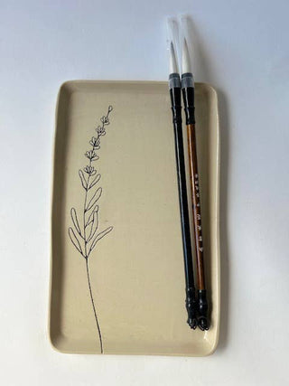 A beige rectangular ceramic plate with a minimalist black line drawing of a plant, holding two black chopsticks with white handles and a brown handle.