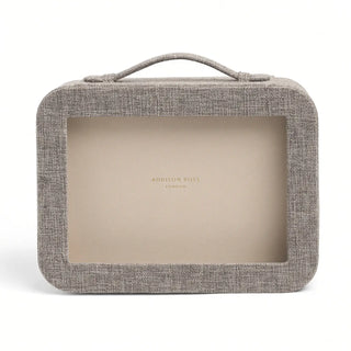A rectangular Addison Ross London cosmetic case with a textured gray linen exterior, a top handle, and a clear front window showing a beige interior. The Addison Ross London brand name is printed in gold on the interior.