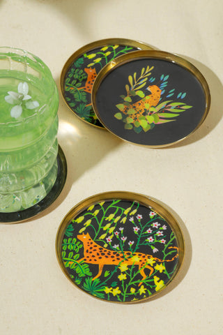 Three round coasters with gold-toned metal rims and a black background, decorated with colorful botanical and animal illustrations, arranged on a light beige surface next to a green glass tumbler.