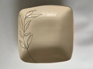 A square ceramic plate with rounded corners in a warm beige color, featuring a delicate black line drawing of a leafy branch on the left side, set against a neutral gray background.