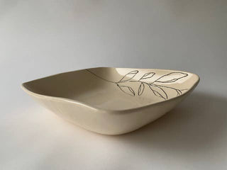 A square ceramic bowl with rounded organic edges, in a warm cream-beige glaze, decorated with a delicate hand-drawn black leaf pattern on the inside, set against a neutral light gray background.