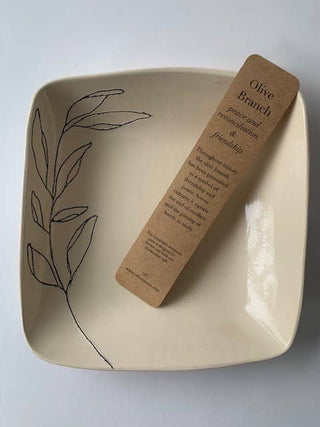 A square beige ceramic bowl with a black line-art olive branch design on the left side, containing a brown kraft paper bookmark labeled 'Olive Branch peace branch and reconciliation, & friendship'.