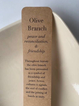 A rectangular kraft paper bookmark or card with rounded corners printed with the text 'Olive Branch peace and reconciliation, & friendship' and a short quote below, photographed leaning against a white ceramic dish.